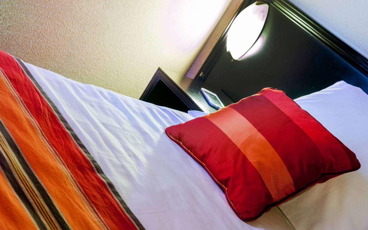 Lit d'une chambre individuelle avec lampe et coussin rouge, hebergement occitanie, Hôtel Saint-Sauveur. 